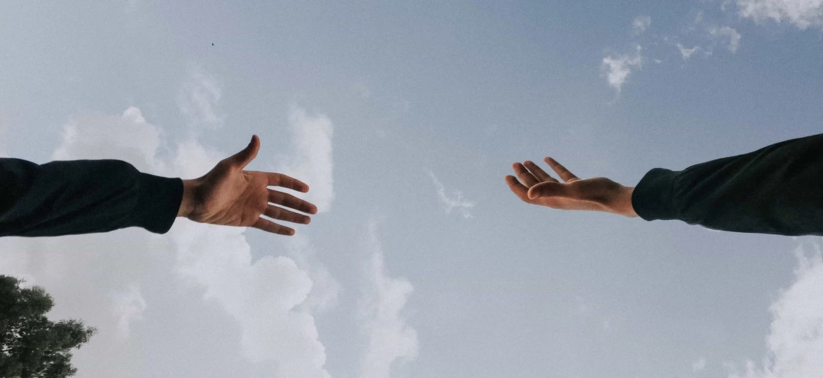 Shot from below, a picture of hands reaching towards each other with the backdrop of a partly cloudy daytime sky